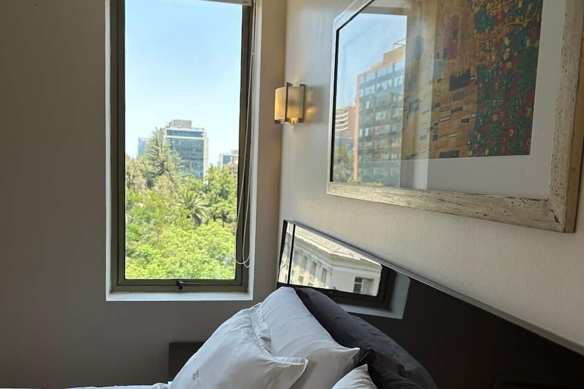 A hotel room with a bed, a window with a view of buildings and trees, a wall-mounted light, and a framed picture.