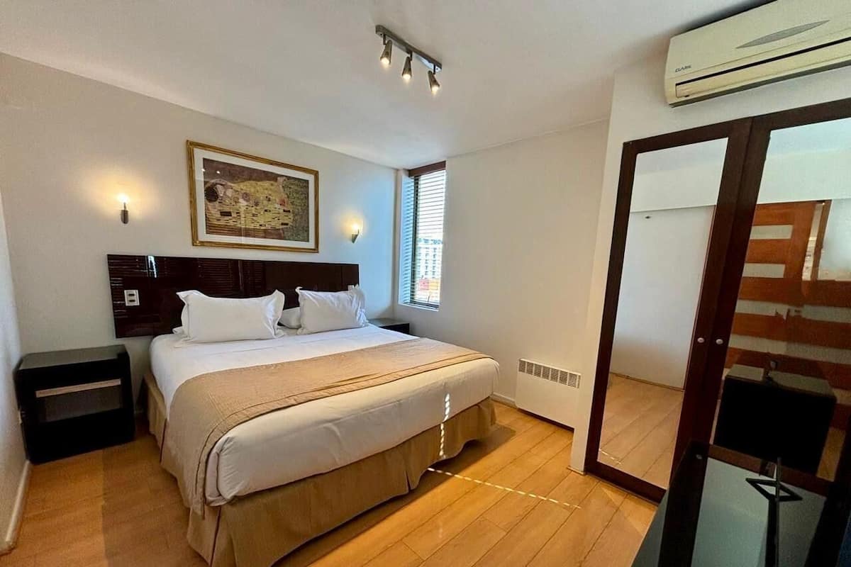 A bedroom with a bed, bedside tables, a wall-mounted air conditioner, and a window with blinds.
