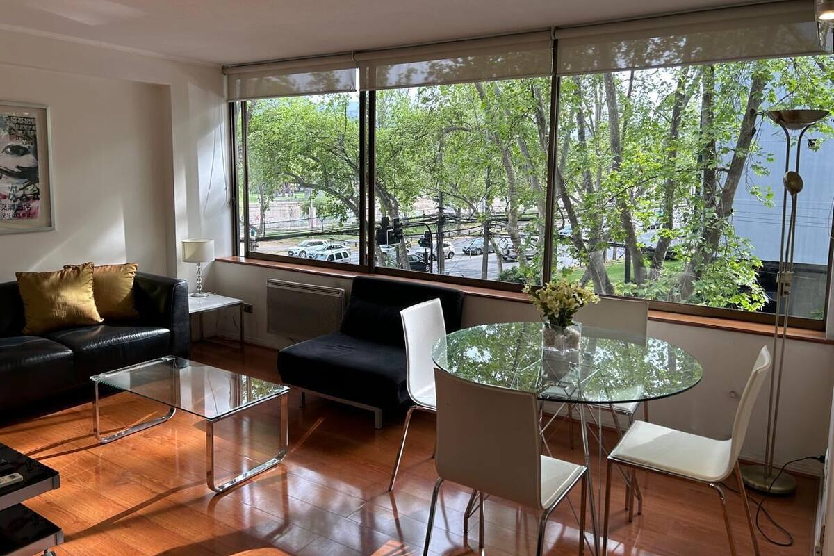 A modern living room with a sofa, a glass coffee table, and a round table with chairs. Large windows offer a view of trees and a street.