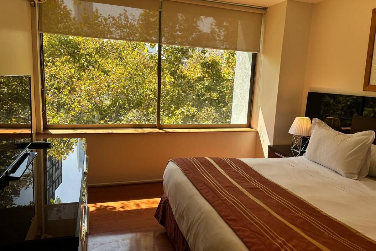 A hotel room with a large window, a bed with a striped bedspread, a bedside table with a lamp, and a desk with a chair.