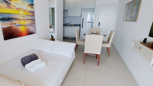 Una habitación de hotel pequeña con una cama, una mesa de comedor con sillas, una zona de cocina y un cuadro de una puesta de sol.