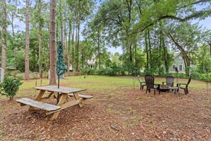 A picnic area with a wooden table and chairs, surrounded by trees and a house in the background.