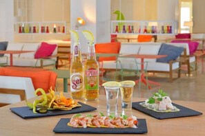 Una mesa de comedor con una variedad de platos, incluyendo camarones y un postre, en un moderno entorno hotelero con sillas color naranja y un interior luminoso.