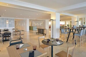 Een moderne eetruimte van een hotel met meerdere tafels en stoelen, een bar en een zichtbare keuken op de achtergrond.