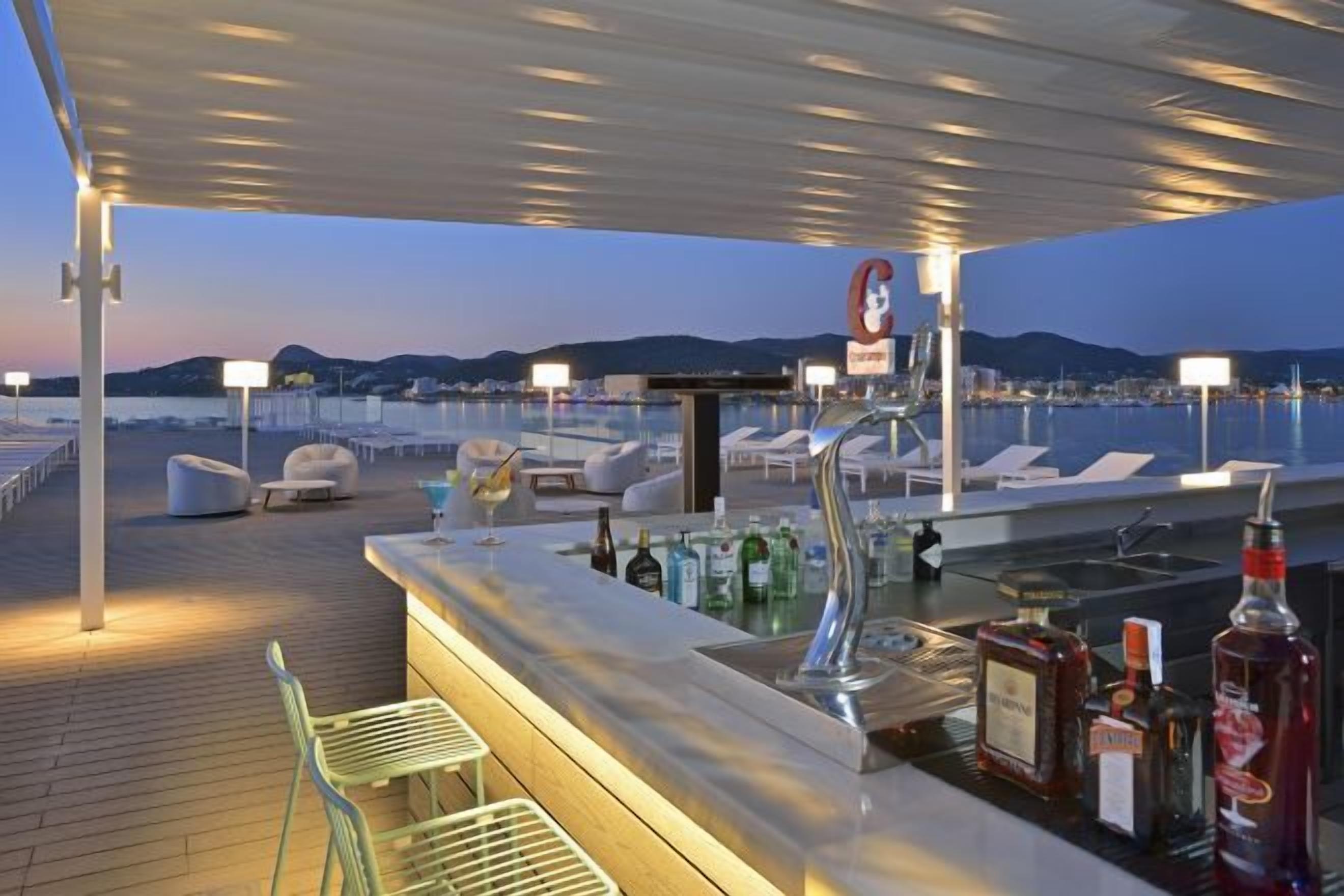 Zona de bar con vista al mar, mesas al aire libre y barra con botellas.
