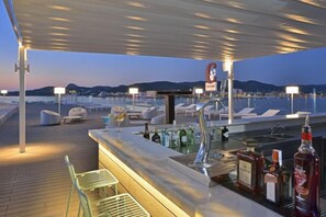 Zona de bar con vista al mar, mesas al aire libre y barra con botellas.