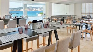 A modern dining area with tables and chairs, large windows with a view of the sea and buildings, and a casual seating arrangement. - Innside by Meliá Ibiza (Ibiza)