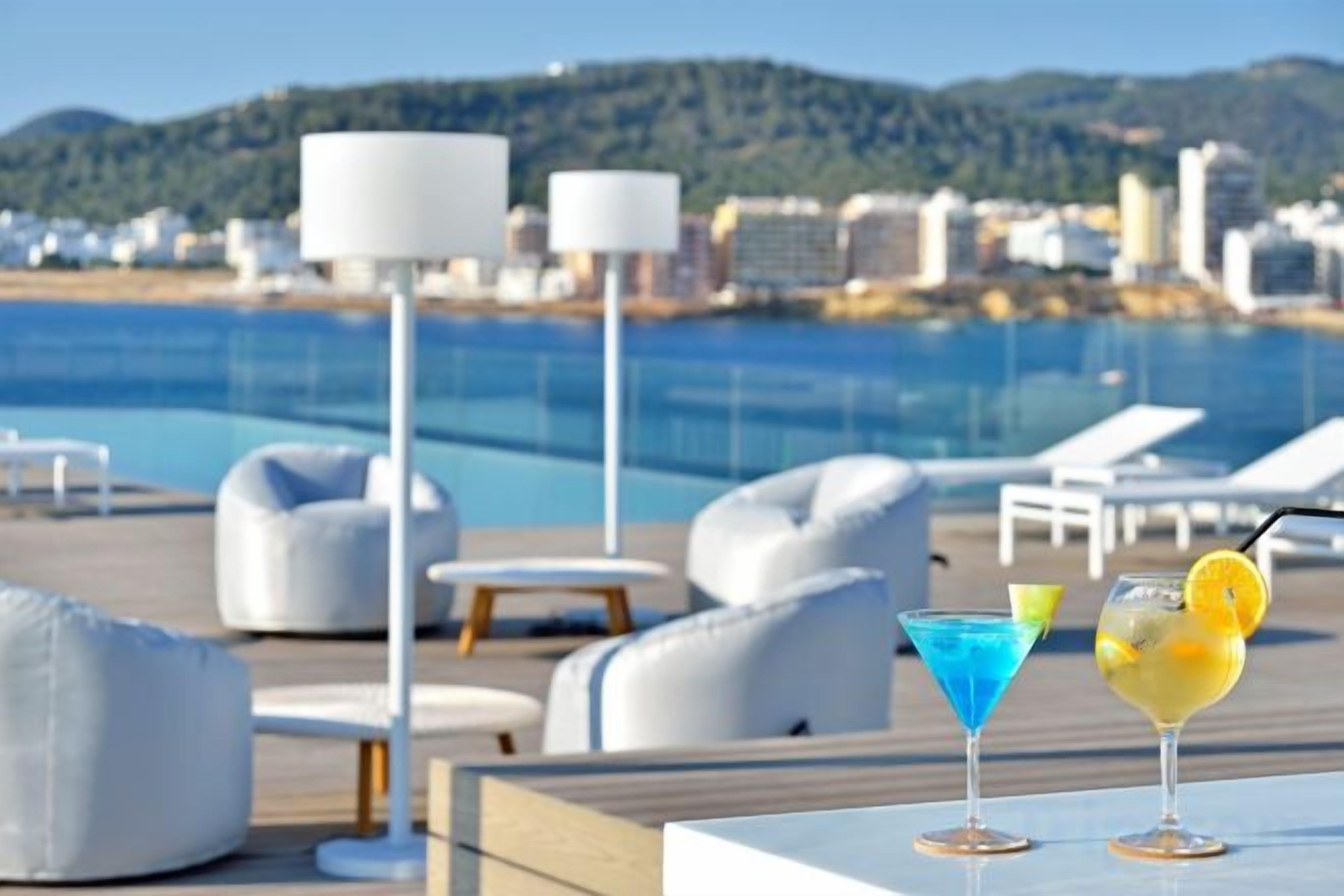 A poolside area with lounge chairs and tables, a pool, and a cityscape in the background.