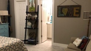Une chambre à coucher comprenant un lit, une commode, une étagère avec des objets décoratifs, un panneau fixé au mur et une porte donnant sur une autre pièce.