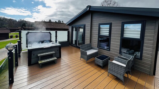 A deck with a hot tub, wicker furniture, and a modern house with large windows. - Modern, spacious lodge at Hunter's Quay Holiday Park. (Dunoon)