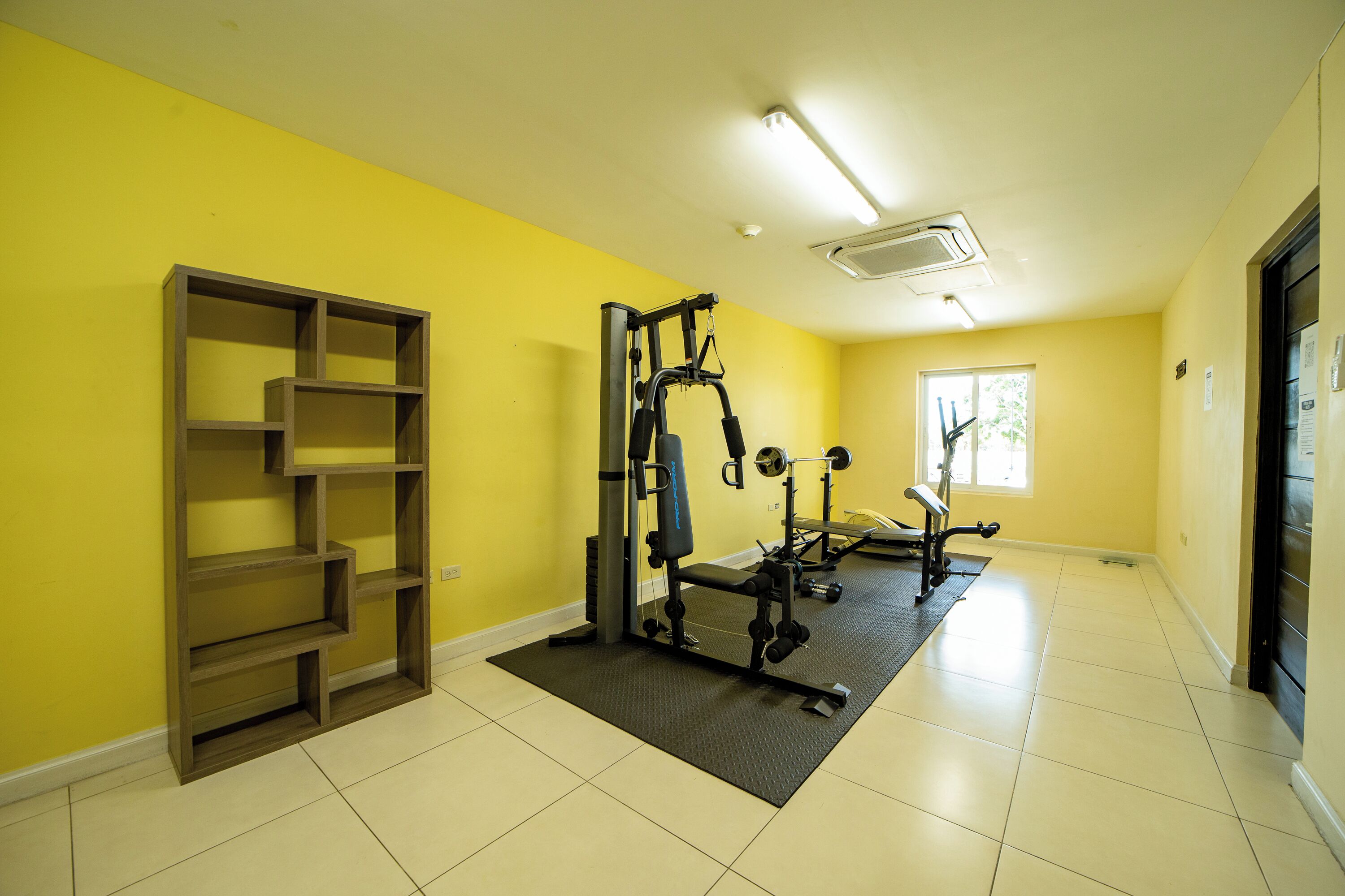 Une salle de sport équipée de matériel d'exercice, d'une étagère et d'une fenêtre laissant entrer la lumière naturelle.
