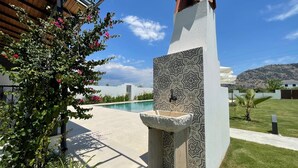 Zona de piscina con pared de azulejos estampados, una fuente de piedra y un exuberante jardín con flores rosadas.