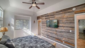 Ein Schlafzimmer mit einem Bett, einem Deckenventilator, einem an einer Holzwand montierten Fernseher und einem Fenster mit Jalousien.