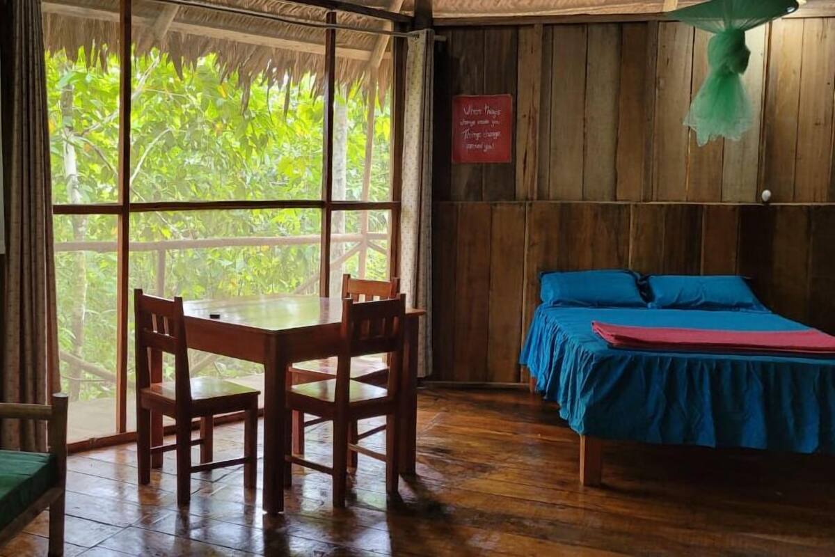 Una habitación con piso de madera, una cama con ropa de cama azul, una mesa de madera con sillas y un ventanal con vistas a la vegetación.