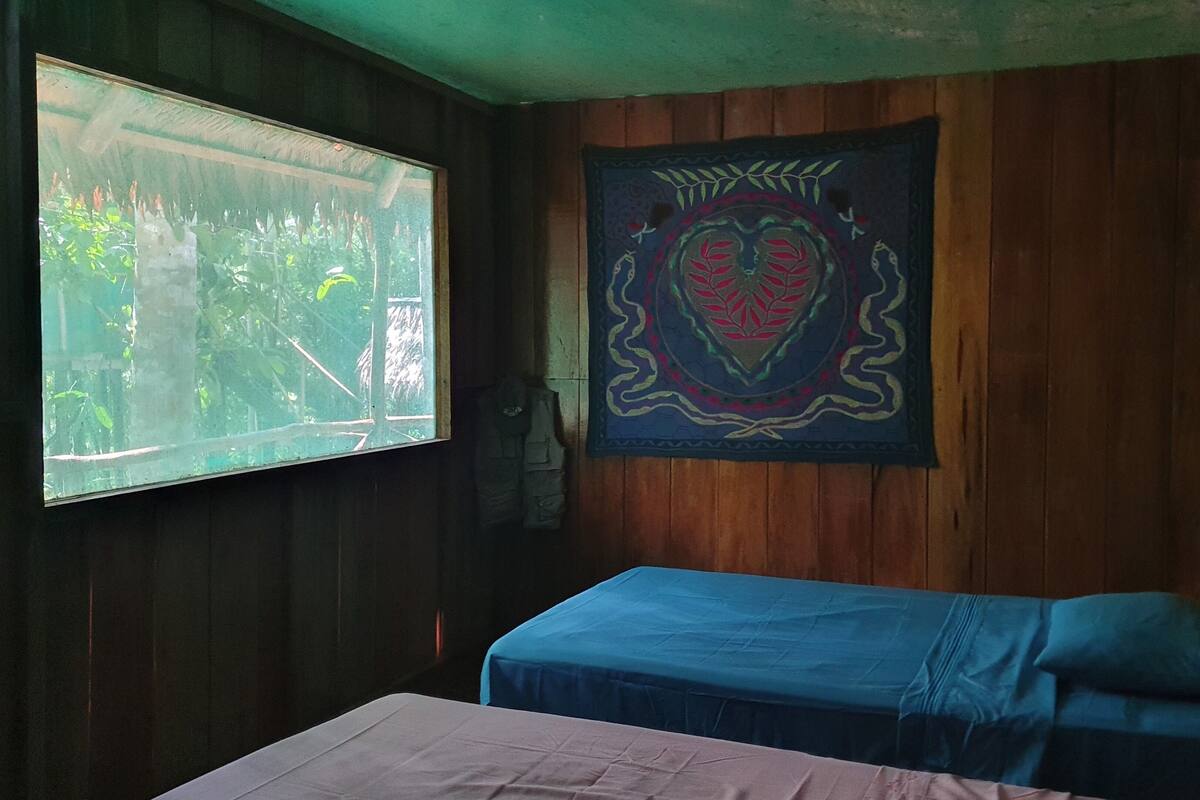 Una habitación con paredes de madera, una ventana que ofrece vistas a zonas verdes, una cama con colcha azul y un tapiz con diseño de corazón.