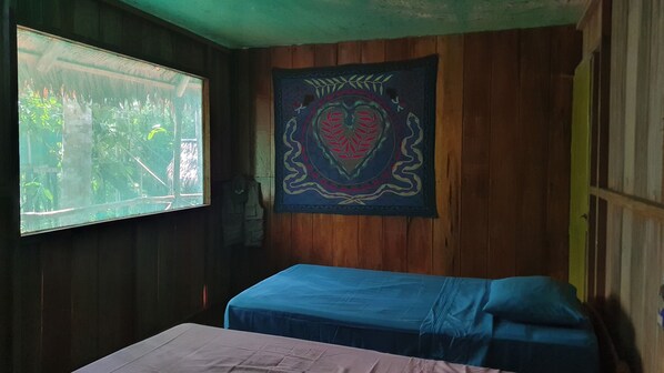 A room with wooden walls, a window with a view of greenery, a bed with a blue bedspread, and a tapestry with a heart design. - Amaca Eco Station (San Juan Bautista)