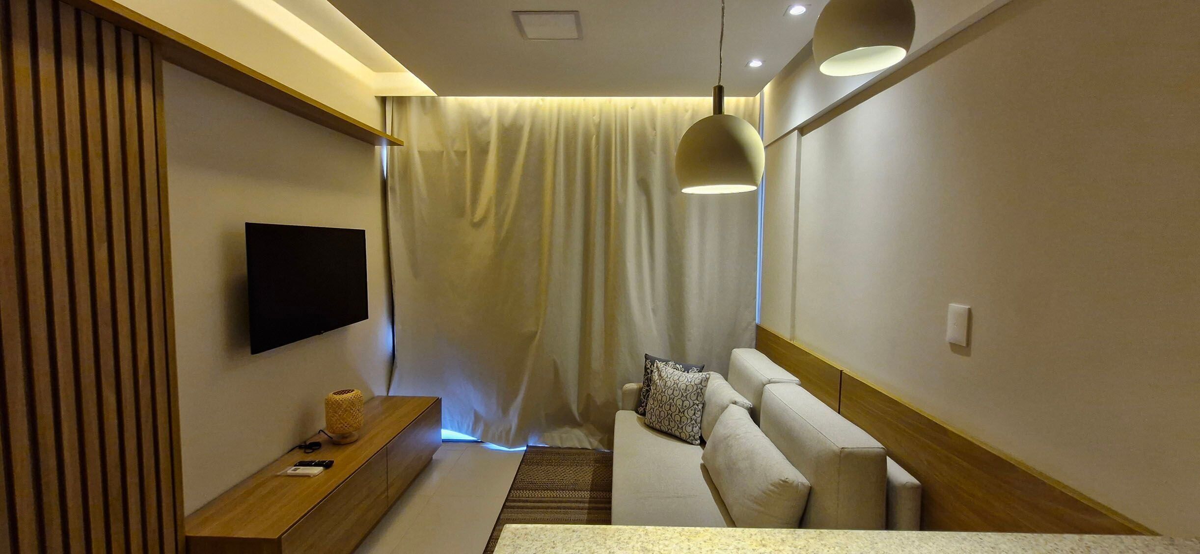 A modern hotel room with a flat-screen TV, a wooden sideboard, a sofa with patterned cushions, and a large window with curtains.