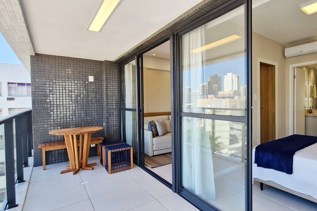 A modern hotel room with a balcony, a bed, a seating area, and a view of the city.