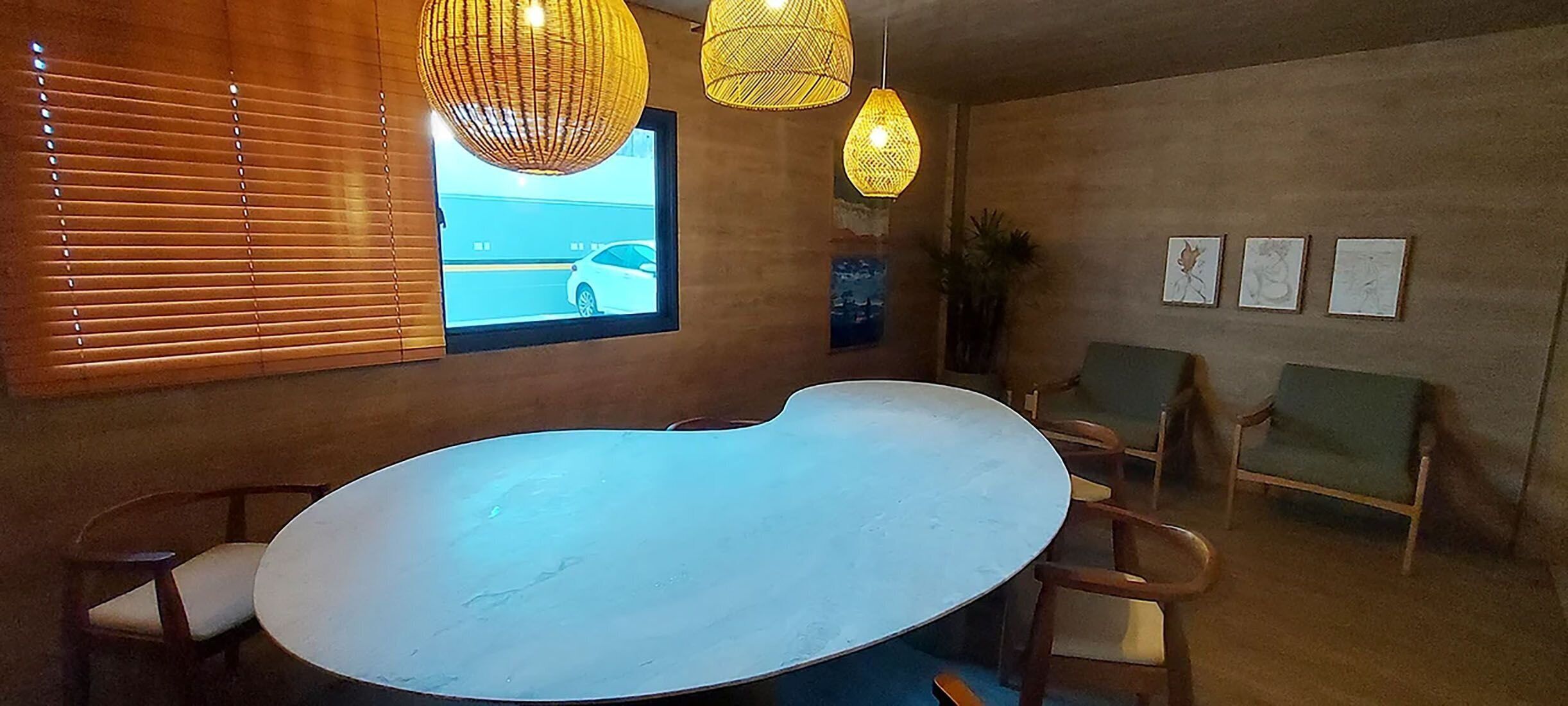 A room with a large oval-shaped white table, wooden chairs, wicker pendant lights, a window with blinds, and framed artwork on the wall.