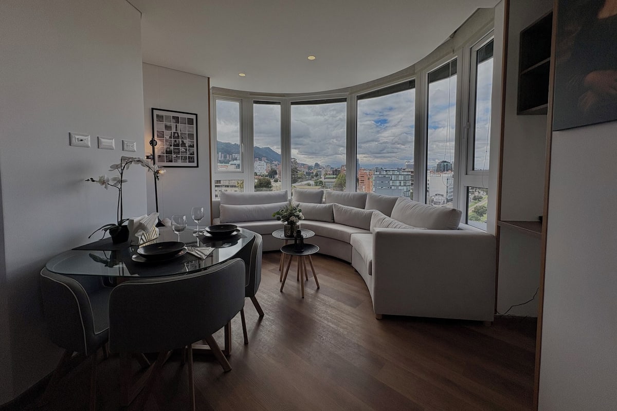 Una sala moderna con un sofá curvo, una mesa de comedor con sillas y grandes ventanas con vista a la ciudad.