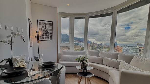 A modern living room with a dining area, large windows with a city view, and a curved sofa. - VOU by Wellcome (Bogotá)