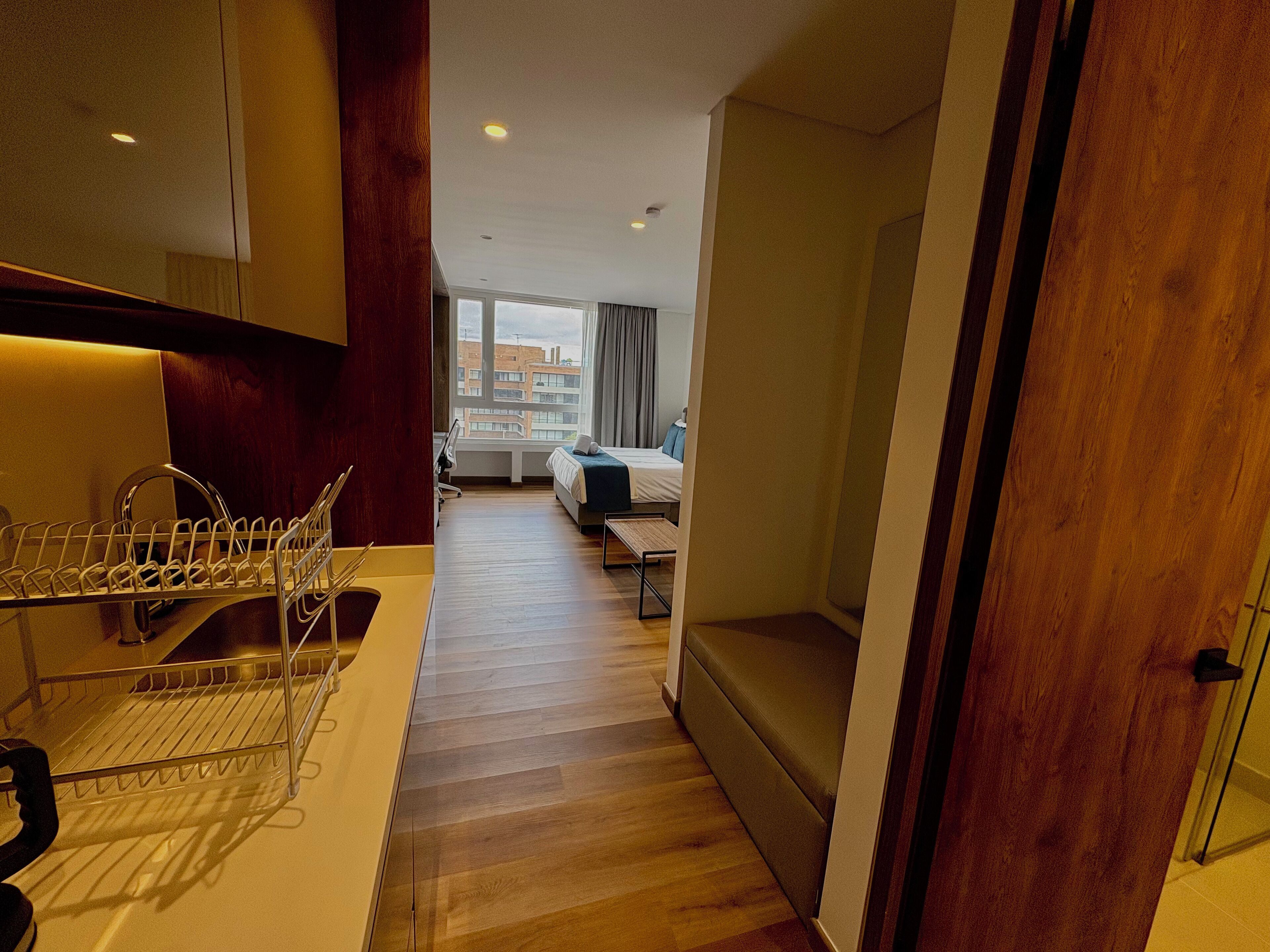 Habitación de hotel con piso de madera, cama con manta azul, escritorio con silla y ventana con vista a edificios.