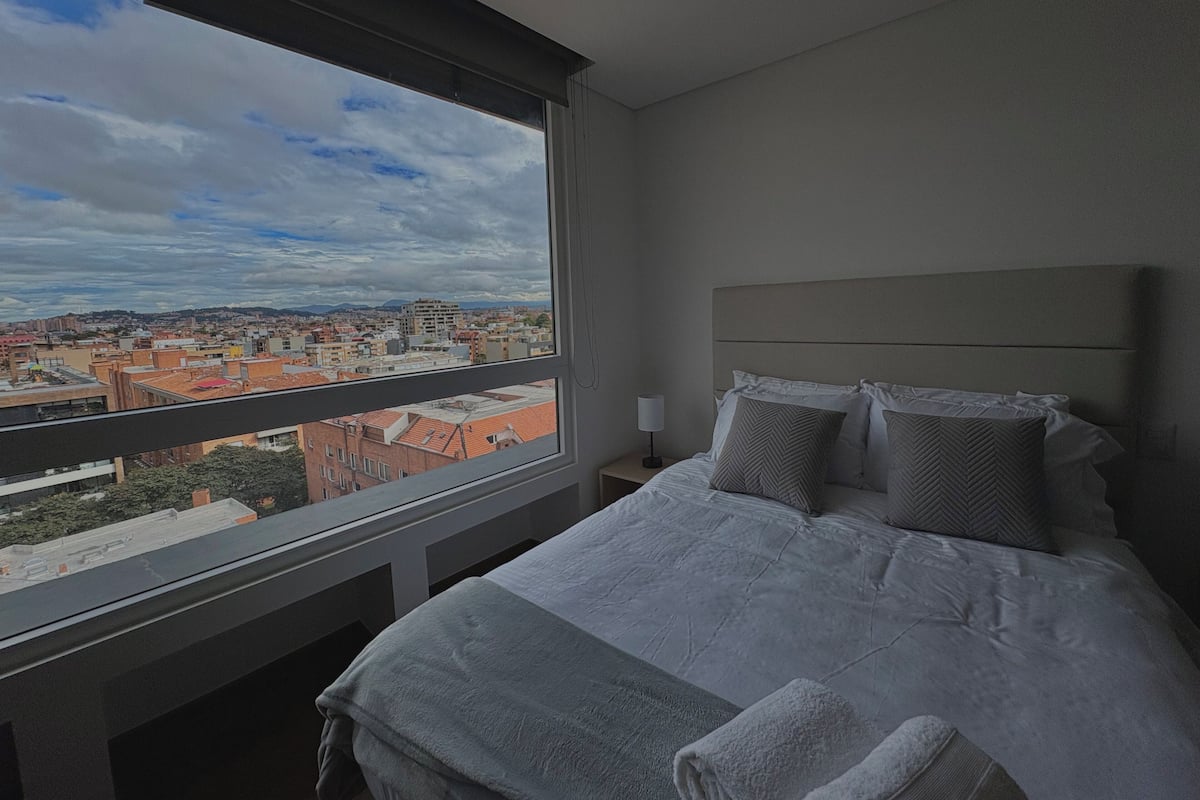 Habitación de hotel con un ventanal amplio que ofrece vistas a la ciudad, una cama bien hecha con almohadas y una mesita de noche con lámpara.