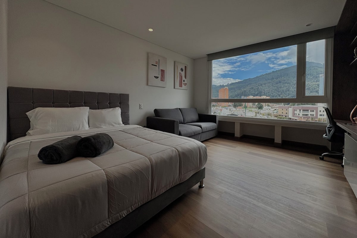 Habitación de hotel moderna con una cama grande, un sofá y un escritorio. Se ve una ventana con vistas a la ciudad.