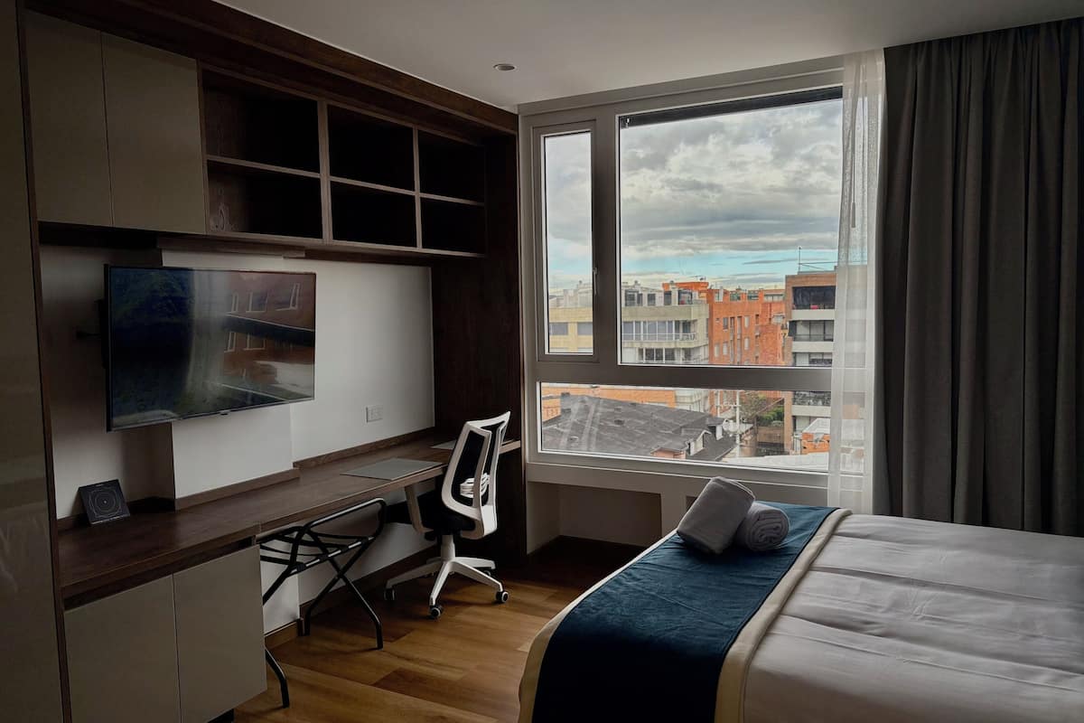 Una habitación de hotel moderna con un escritorio de madera, un televisor de pantalla plana, un sillón cómodo, una cama con cubrecama azul y un ventanal con vistas a la ciudad.