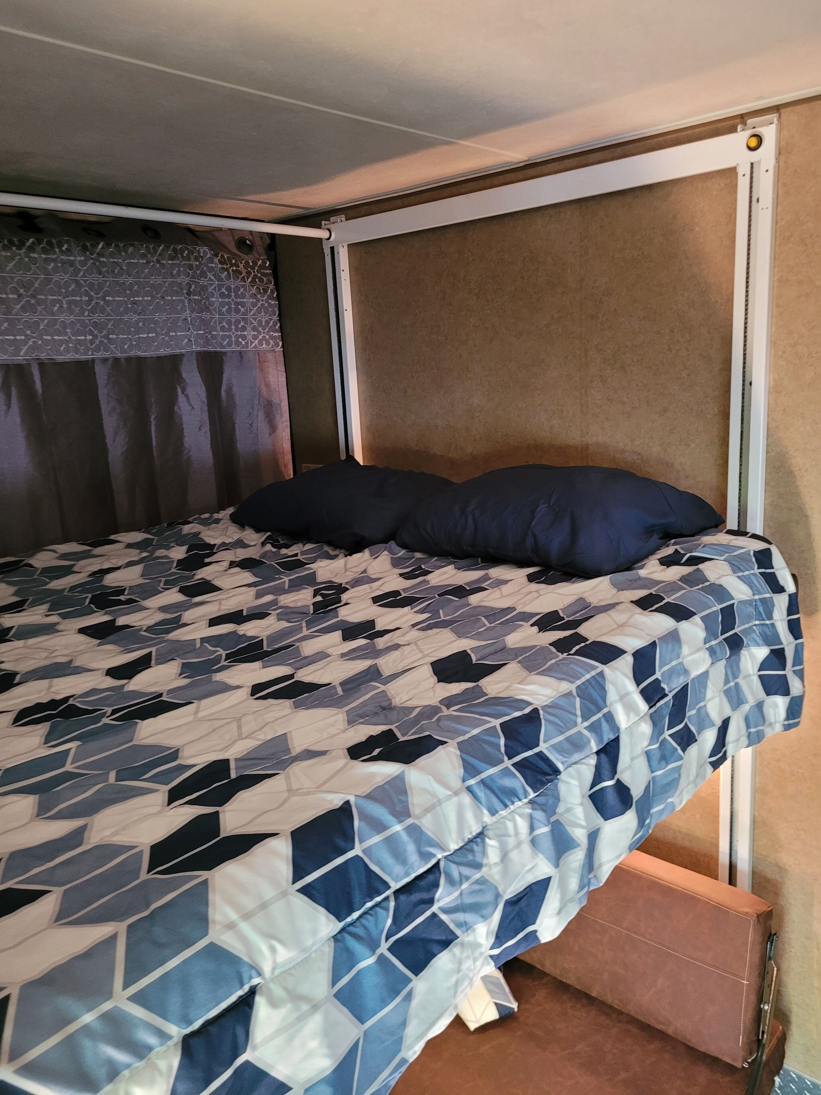 A bed with a patterned bedspread, a dark pillow, and a suitcase under the bed.