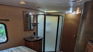 A compact RV bathroom with a shower, sink, and a bed with patterned bedding. - 2-bedroom RV in beautiful Bowser with WiFi, AC (Bowser)
