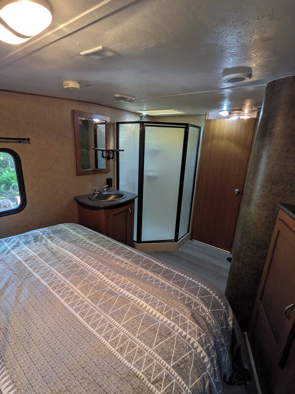 Ein kleines Badezimmer in einem Wohnmobil, ausgestattet mit Dusche, Waschbecken und einem Bett mit gemusterten Betttextilien.