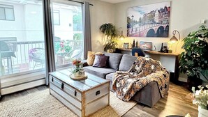 A living room with a sofa, a coffee table, a TV, a bookshelf, and a window with curtains. - Downtown Condo near Rogers | Parking, Pool, Gym (Edmonton)