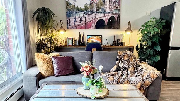 A living room with a vintage suitcase coffee table, a sofa, a painting of a bridge, and a potted plant. - Downtown Condo near Rogers | Parking, Pool, Gym (Edmonton)