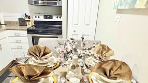 A dining table set with gold and white tableware, a vase with flowers, and a kitchen in the background. - Downtown Condo near Rogers | Parking, Pool, Gym (Edmonton)