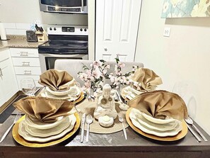 A dining table set with gold and white tableware, a vase with flowers, and a kitchen in the background. - Downtown Condo near Rogers | Parking, Pool, Gym (Edmonton)