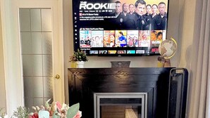 A living room with a TV displaying a show, a fireplace, and decorative plants. - Downtown Condo near Rogers | Parking, Pool, Gym (Edmonton)