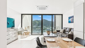 Een moderne woonkamer met een eethoek, grote ramen met uitzicht op een kustplaats en een flatscreen televisie op een houten kast.