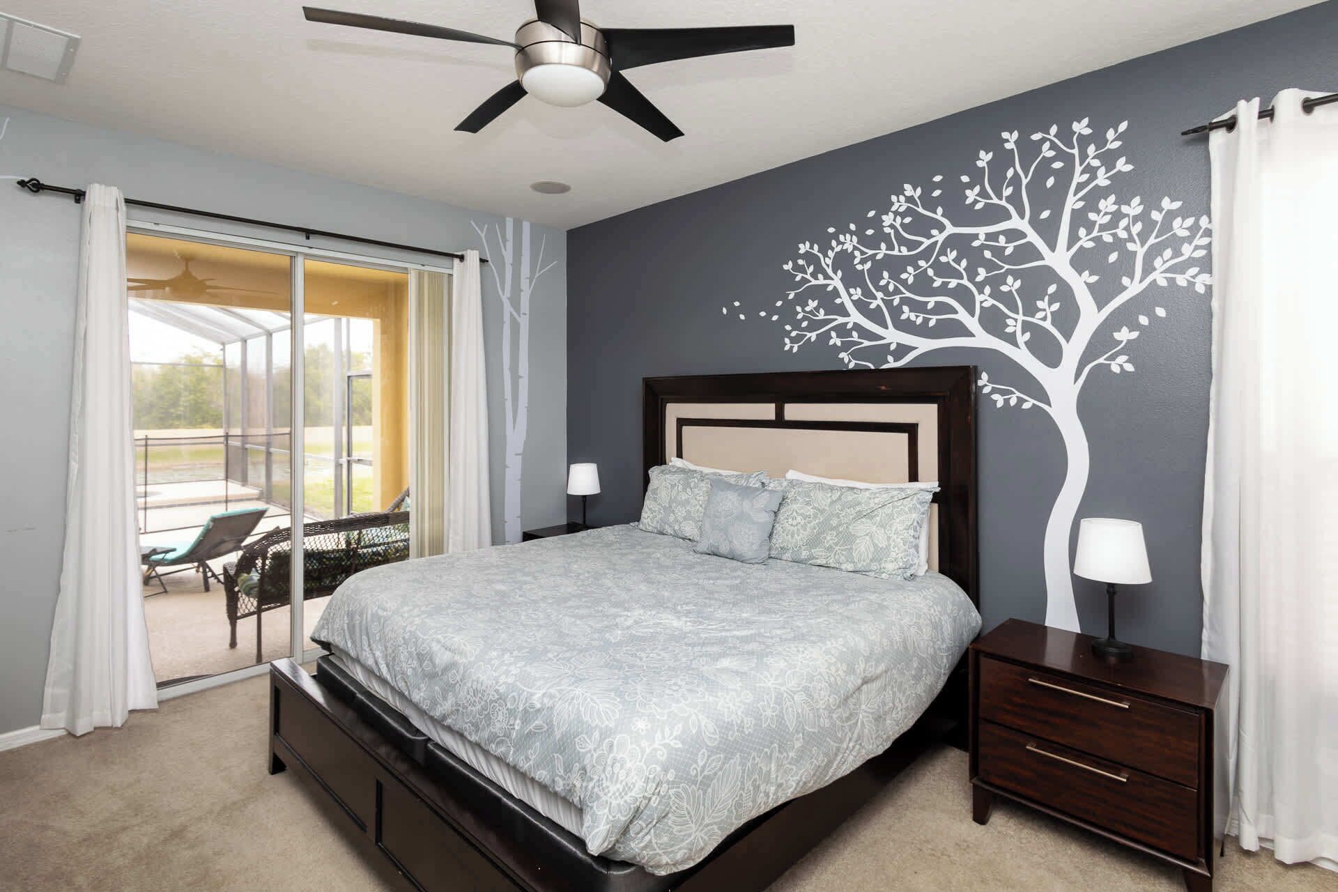 Un dormitorio con cama, mesita de noche, ventilador de techo y un mural de un árbol en la pared.