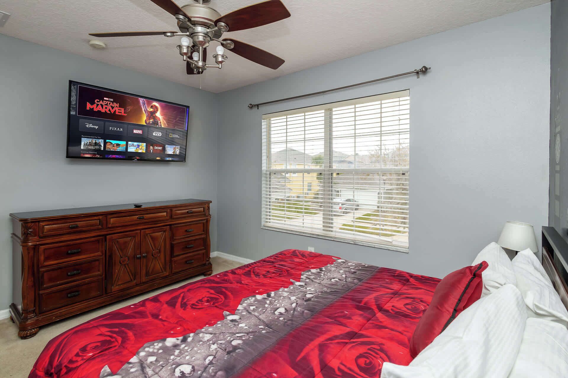 A bedroom with a bed, a dresser, a ceiling fan, a TV on the wall, and a window with blinds.