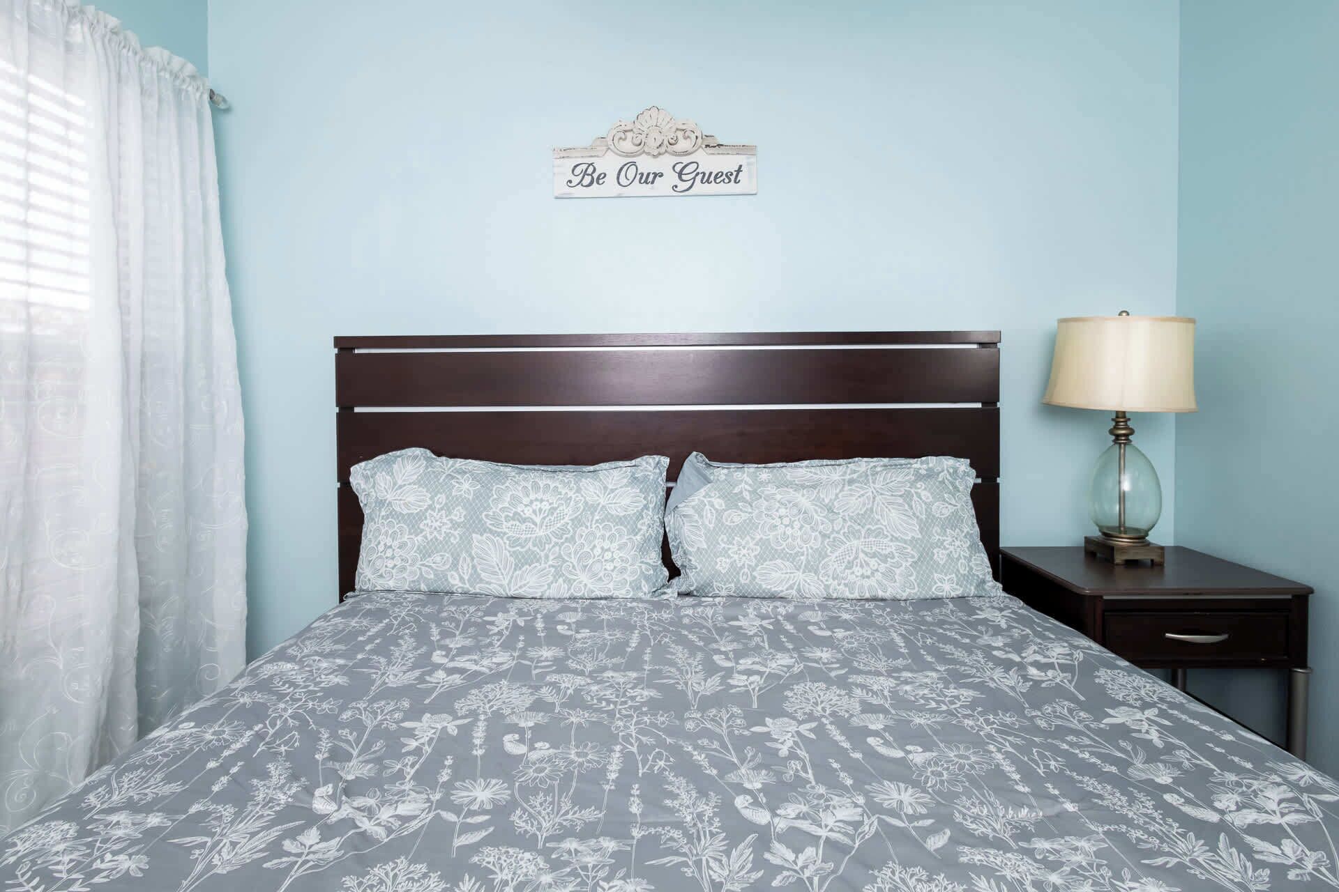A bed with a floral-patterned bedspread, a dark wooden headboard, and a sign above the bed that reads 'Be Our Guest.'