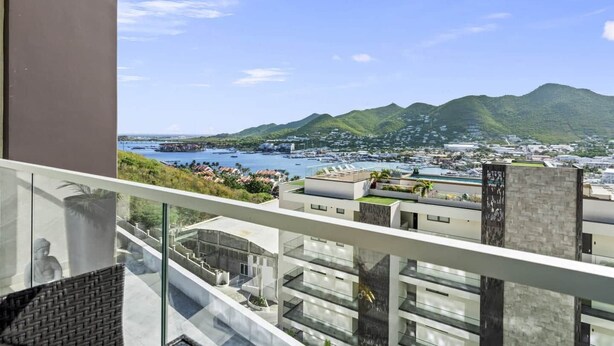 A modern building with a glass balcony overlooking a coastal cityscape with mountains in the background.