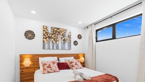 Quarto moderno com cabeceira de madeira, cama grande com várias almofadas, obra de arte na parede e janela com vista.