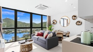 Sala de estar moderna com uma janela ampla que oferece vista para um corpo de água e colinas, um sofá cinza com almofadas, uma mesa de centro redonda de madeira e uma área de cozinha com uma chaleira.