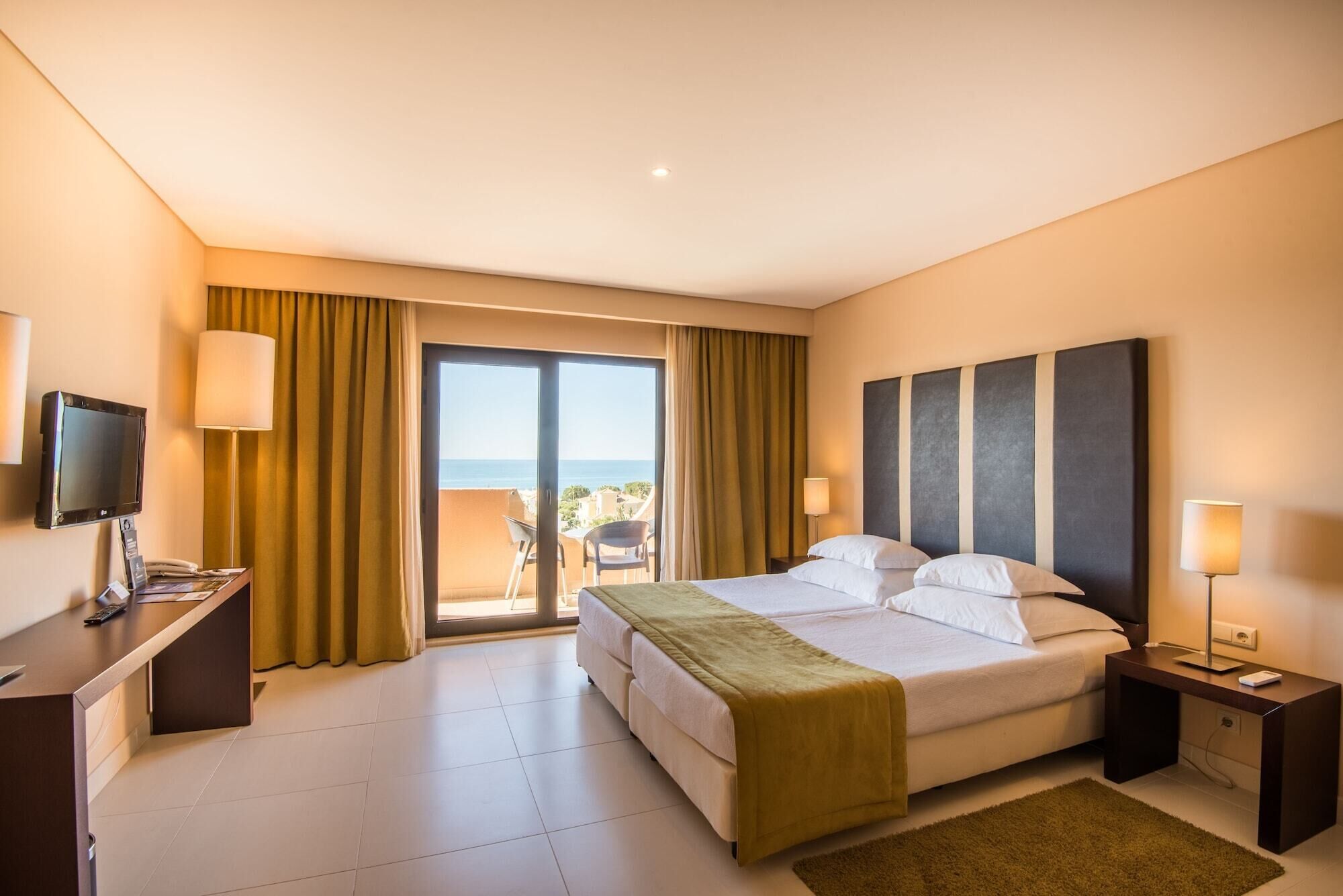 A hotel room with a bed, a desk, a TV, a balcony with a view of the sea, and a sliding door.