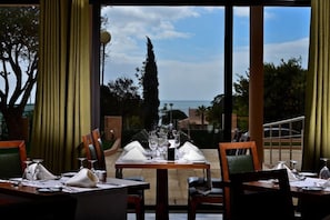 Mesa de comedor lista para dos con vista al mar, cortinas y una lámpara.