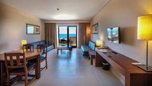 Ein Hotelzimmer mit Esstisch, Stühlen, einer Sitzecke, einem Fernseher und Blick aufs Meer durch das Fenster.