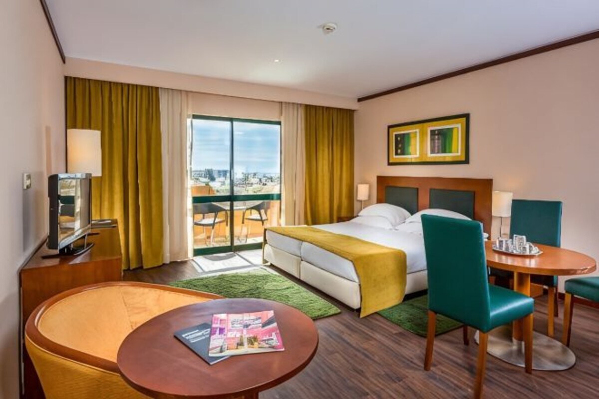 A hotel room with a bed, a desk, a chair, a table, and a view of the city through a large window.