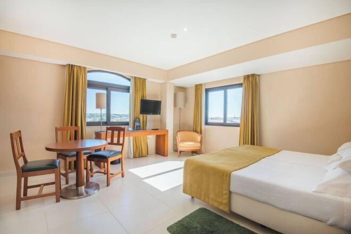 Una habitación de hotel con cama, escritorio con silla, televisor y ventanas amplias.