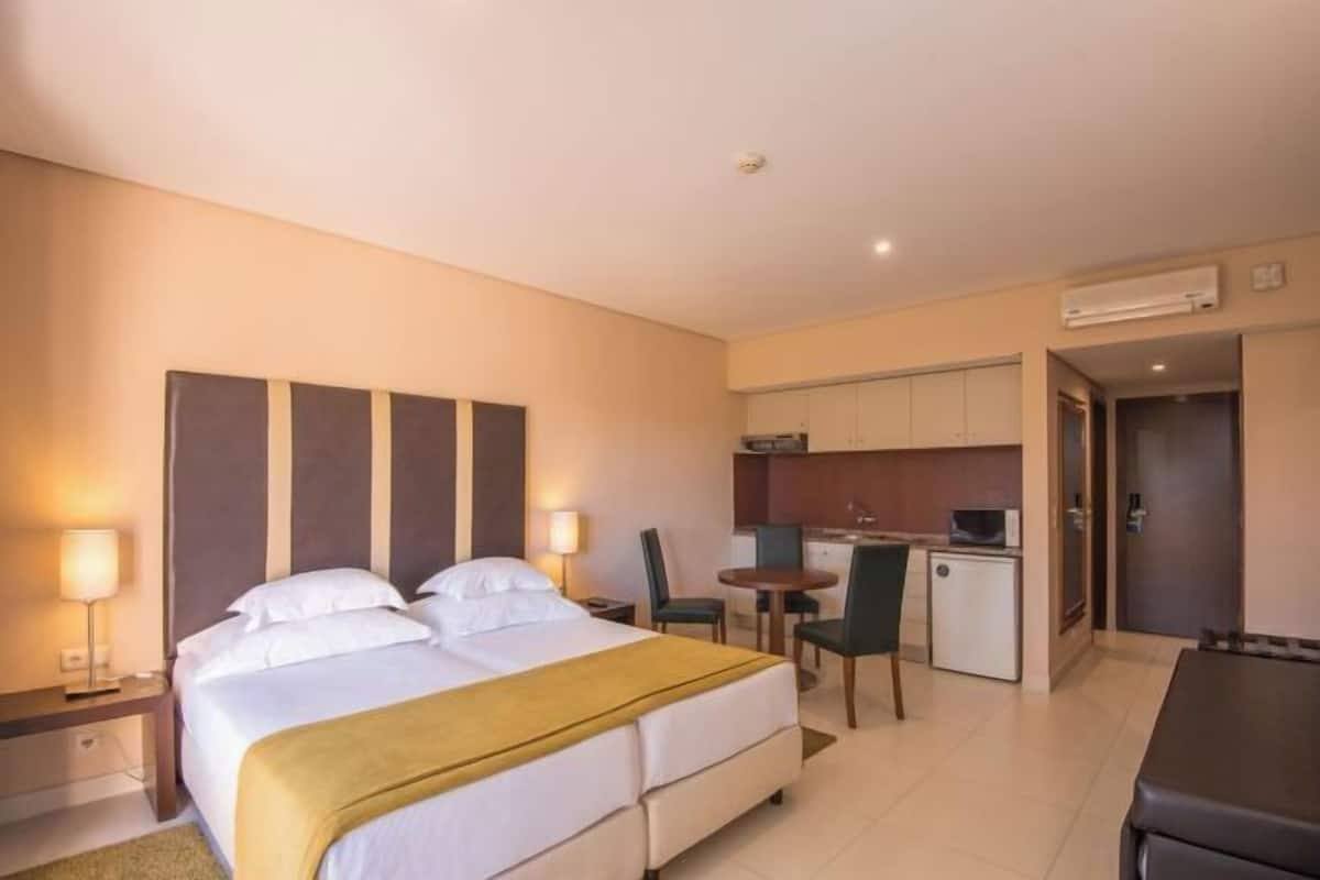Habitación de hotel con cama, mesitas de noche, escritorio, silla, kitchenette y unidad de aire acondicionado.